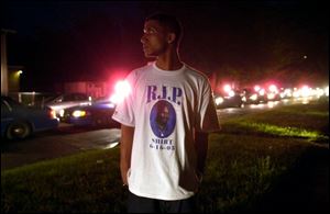 Jomo Brooks, 27, watches police cars parade past with lights and sirens on to signal the start of a curfew. He wears a shirt with an image of the motorcyclist whose death sparked the riots.