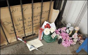 Candles, flowers, and sympathy cards are set up in a make-shift memorial outside the front door of Tamara's Carryout on Columbus Street at Erie Street. The cardboard sign expresses sympathy and prayers from the neighborhood.