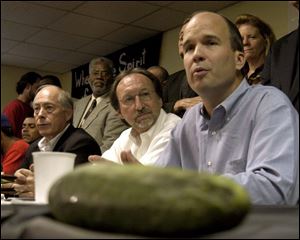 With a cucumber lying on the table, Bill Bryan, president of Mt. Olive Pickle Co., talks to reporters; at left, is FLOC president Baldemar Velasquez; in the center is Stan Eury, of the growers' group.