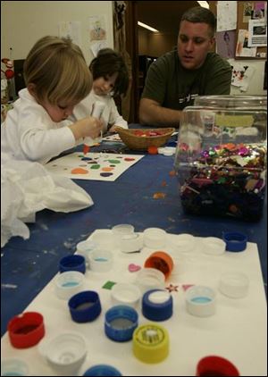 Perrysburg residents Grace Burkin, 2, left, and her sister Hope, 5, create bottle-cap art at the Toledo Museum of Art's Family Center while their father, Donny, offers helpful tips.