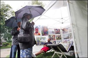 Anthony Whiting, left, and his wife, Minnie Whiting, center, talk with local artist Aaron Bivins about his paintings while he takes cover from the rain in his exhibition booth at the Toledo Botanical Garden.