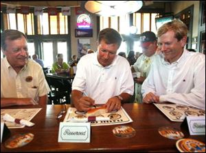 James Seney, left, former mayor of Sylvania, and Jon Stainbrook, right, Lucas County GOP chairman, watch John Kasich as the candidate for Ohio governor signs a hot dog bun at Packo's in the Park.