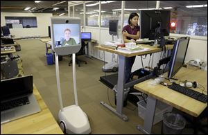 Senior software engineer Josh Faust, seen on screen, navigates his company's office as fellow engineer Stephanie Lee works on a project. The roving computer screen, with video cameras, microphones, and speakers, rides on motorized wheels.