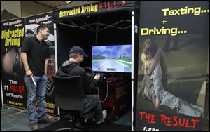 Owens student Alex Bohland texts and drives in the simulator, as  Adam Kelly, an employee of the firm which tours the simulator, watches.