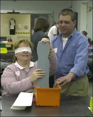 Dr. Beth Rohrbacher, director of pupil personnel in Perrysburg, works blindfolded to fit a sock onto a rack so they are outside in. With her is Todd Lashuay, production specialist.