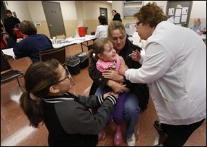 Bridgett Rys holds her daughter, Isabella, 3, while she gets her shot from nurse Cindy McLeod at the Toledo-Lucas County Health Department. Anabella Rys offers support by holding her sister’s hand.