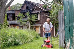 Tim Johnson mows a strip of yard next to his home in the 2000 block of Broadway in South Toledo. The house behind his home at 925 Harding Dr., in the background, caught fire more than five years ago and is still standing, to his frustration, and a house that once stood beside his house was torn down after a fire, but the grass is tall.